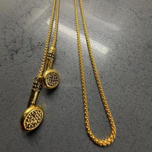 May include: A gold-toned necklace featuring a delicate box chain and two dangling, circular pendants. Each pendant has a textured, granulated surface with a raised border, suspended from the chain by a slender, cylindrical metal connector piece.