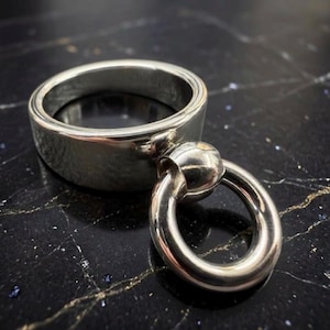 May include: A polished silver-tone metal ring featuring a wide, smooth band with a smaller, circular ring charm attached to the front by a rounded connector. The metal has a reflective, high-shine finish throughout the entire piece.