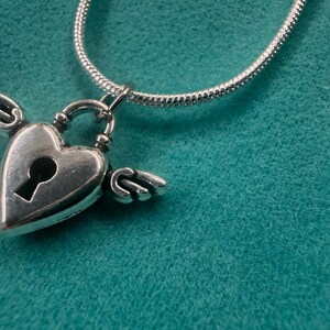 May include: A silver heart-shaped pendant with small wings and a keyhole, hanging from a silver chain. The pendant is set against a teal textured backdrop. A romantic necklace.