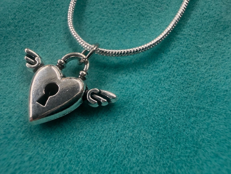 May include: A silver heart-shaped pendant with small wings and a keyhole, hanging from a silver chain. The pendant is set against a teal textured backdrop. A romantic necklace.