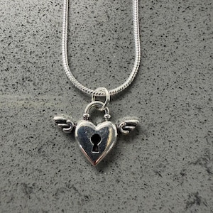 May include: A silver necklace with a heart-shaped pendant, complete with a keyhole and small wings. The pendant is suspended from a fine snake chain. The jewellery is set against a grey background.
