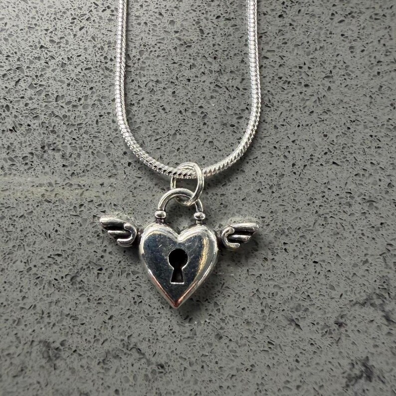 May include: A silver necklace with a heart-shaped pendant, complete with a keyhole and small wings. The pendant is suspended from a fine snake chain. The jewellery is set against a grey background.