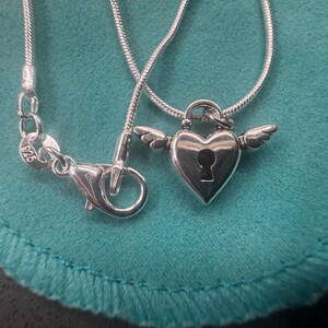 May include: A silver necklace featuring a heart-shaped pendant with a keyhole design and small wings. The necklace has a fine snake chain, and the clasp is visible. The pendant is made of silver.