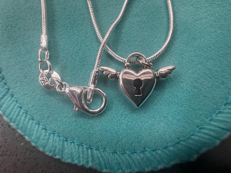 May include: A silver necklace featuring a heart-shaped pendant with a keyhole design and small wings. The necklace has a fine snake chain, and the clasp is visible. The pendant is made of silver.