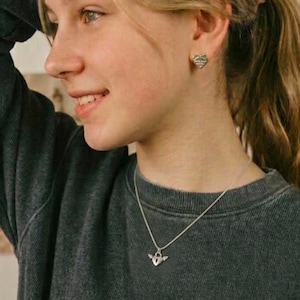 May include: A silver necklace featuring a heart-shaped pendant with wings, complemented by heart-shaped stud earrings. The jewellery is worn with a dark grey sweatshirt.
