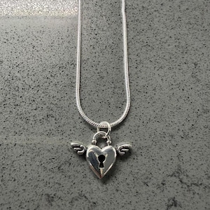 May include: A silver necklace featuring a heart-shaped pendant with a keyhole and wings. The chain is a thin, snake-style design. The pendant is a bright, shiny silver colour.