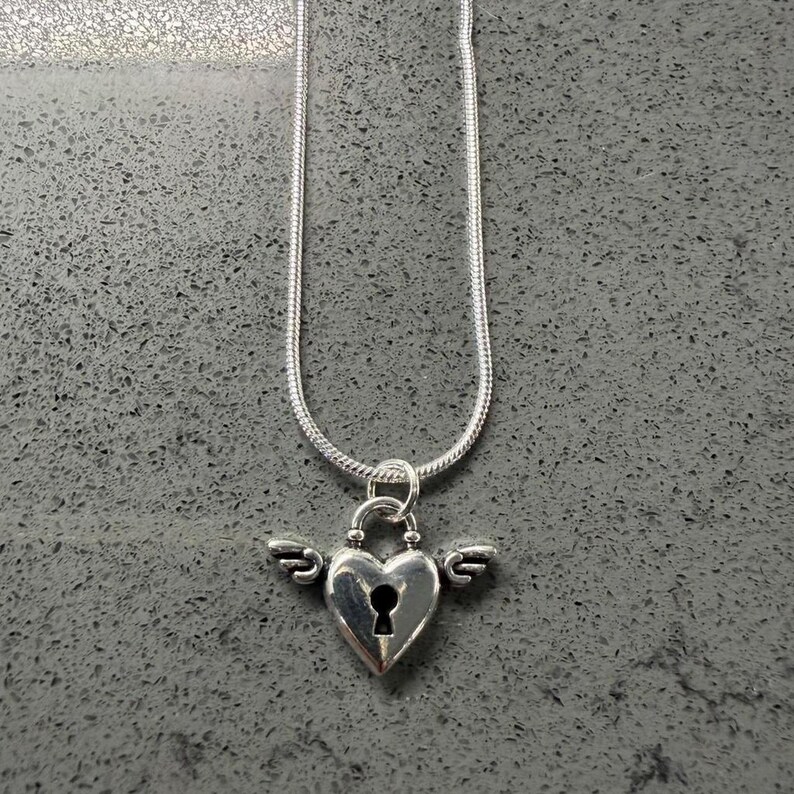 May include: A silver necklace featuring a heart-shaped pendant with a keyhole and wings. The chain is a thin, snake-style design. The pendant is a bright, shiny silver colour.