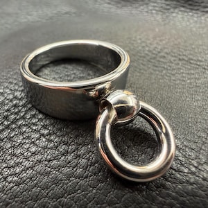 May include: A polished silver-tone metal ring featuring a wide, smooth band with a smaller, circular ring attached to the front by a rounded connector. The metal surface is reflective and smooth, showcasing a clean, metallic finish throughout the piece.