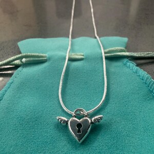 May include: A silver necklace with a heart-shaped pendant, keyhole detail, and small wings. The pendant hangs from a fine chain. The necklace is presented on a teal pouch.