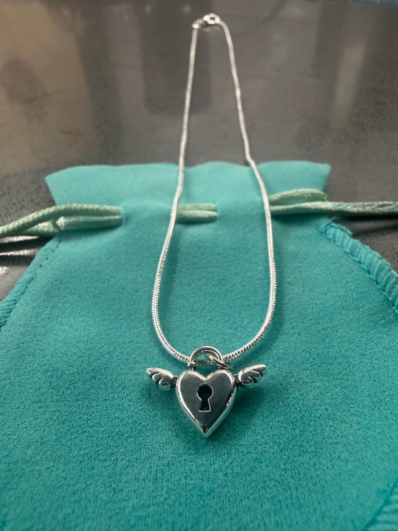 May include: A silver necklace with a heart-shaped pendant, keyhole detail, and small wings. The pendant hangs from a fine chain. The necklace is presented on a teal pouch.
