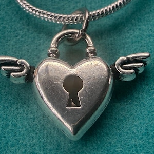 May include: A silver heart-shaped pendant with a keyhole and small wings. The pendant hangs from a silver chain. The heart-shaped lock has a textured surface. The background is teal.