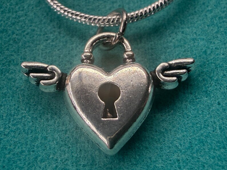 May include: A silver heart-shaped pendant with a keyhole and small wings. The pendant hangs from a silver chain. The heart-shaped lock has a textured surface. The background is teal.