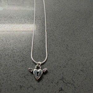 May include: A silver necklace with a heart-shaped pendant, keyhole detail, and small wings. The pendant hangs from a fine chain. The jewellery is set against a dark grey background.