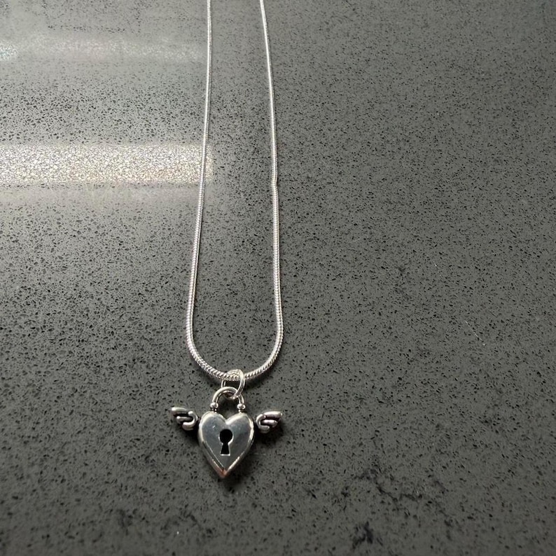 May include: A silver necklace with a heart-shaped pendant, keyhole detail, and small wings. The pendant hangs from a fine chain. The jewellery is set against a dark grey background.