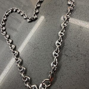 May include: A silver-toned metal chain necklace composed of repeating heart-shaped links. The chain is arranged in a heart shape and features a lobster claw clasp at the bottom, showcasing a polished, reflective surface throughout the entire piece.