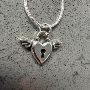 May include: A silver heart-shaped pendant with a keyhole design and small wings, suspended from a silver chain. The pendant is a lock design, and the chain has a snake-like texture. The background is a grey, textured surface.
