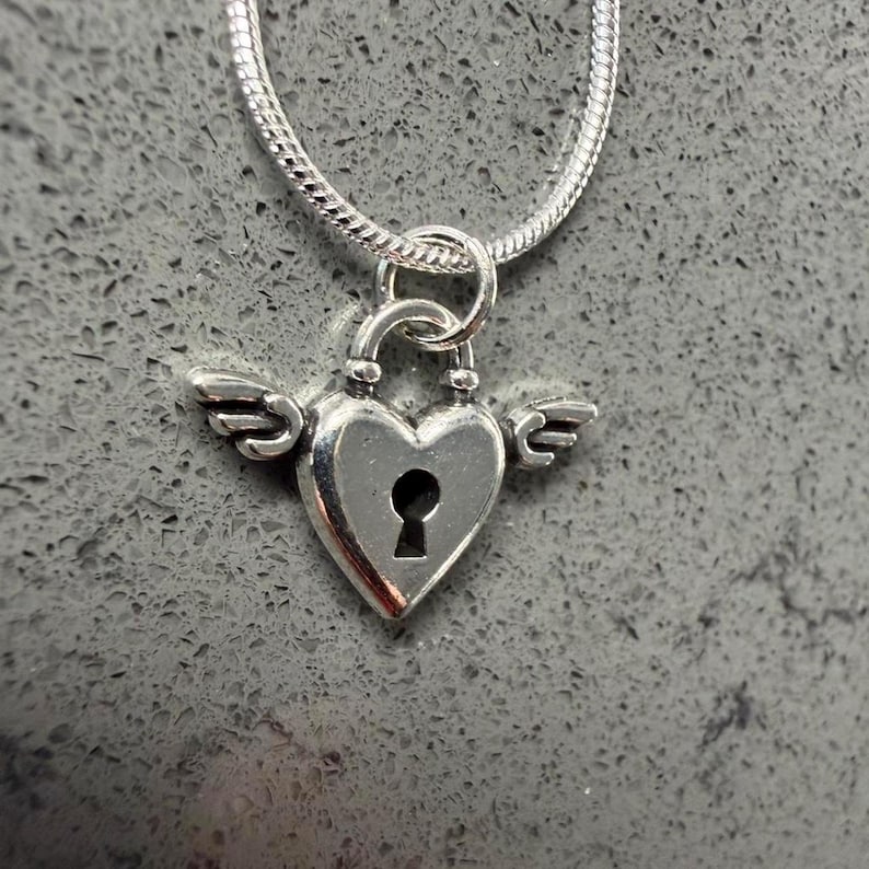 May include: A silver heart-shaped pendant with a keyhole design and small wings, suspended from a silver chain. The pendant is a lock design, and the chain has a snake-like texture. The background is a grey, textured surface.
