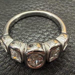 May include: A silver-toned metal ring featuring a central round-cut clear stone set in a bezel, flanked by two rectangular-cut clear stones on each side. The band has a polished finish with textured detailing around the stone settings.