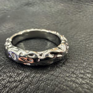 May include: A silver-toned ring featuring a continuous, irregular, and textured surface with small, recessed indentations throughout the band. The metal has a polished, reflective finish that highlights the organic, crater-like pattern across the exterior.