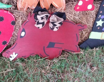 May include: A red wooden pig with a black and white bow, a black eye, and a black stripe on its back. The pig is lying on a bed of straw.