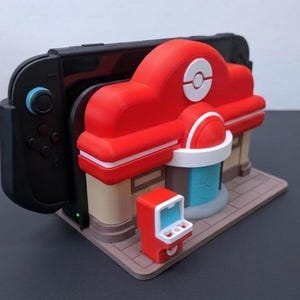 Puede incluir: Un soporte para Nintendo Switch impreso en 3D, rojo y blanco, con forma de edificio, con una pequeña máquina recreativa roja. El soporte sostiene una consola Nintendo Switch negra. El edificio tiene un diseño blanco y rojo con un logotipo circular.