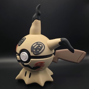 May include: A beige and black figurine featuring a spherical body with a central white button, a pointed top, and a jagged base. The figure has black circular markings on the sides, a brown tail, and a small black mouth detail near the bottom.
