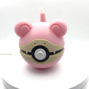 May include: A pink and tan 3D-printed figure resembling a cartoon character. The figure has a spherical body, large ears, and a tail. The mouth area is tan with white teeth, and the center has a black circle with a white button.