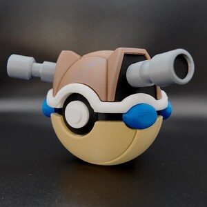 May include: A spherical toy with a tan and brown shell, featuring a white center button, two blue side accents, and two gray cylindrical cannons extending from the top. The object has a layered, 3D-printed texture throughout its surface.