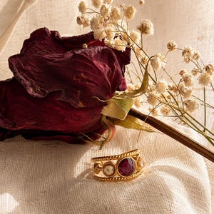 Puede incluir: Un anillo dorado con una piedra central redonda de color rojo intenso, flanqueada por dos piedras redondas más pequeñas de color blanquecino. El anillo tiene un diseño de cuerda trenzada y está sobre un fondo de rosa borgoña seca y flores blancas.