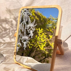 Peut inclure: Miroir rectangulaire avec un cadre en bambou et un motif floral gravé sur le verre. Le miroir est soutenu par un support en bois. Le motif floral est blanc et le cadre est de couleur marron clair.