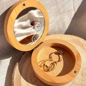 Peut inclure: Une boîte à bijoux ronde en bois avec un couvercle à charnière et un miroir. Le couvercle est ouvert, révélant une petite décoration de coccinelle. Deux bagues en or sont à l'intérieur. La boîte est posée sur une surface en bois.