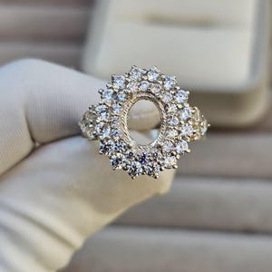 May include: A silver ring with an oval centre setting, encircled by many small, sparkling clear stones. The ring's design includes a halo of stones around the centre, with extra stones on the shoulders, producing a dazzling effect. The ring is shown against a white background.