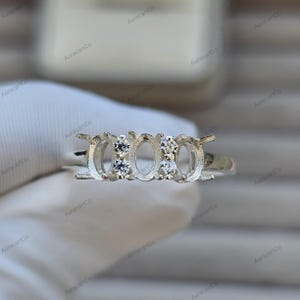 May include: A silver ring setting with three oval spaces for stones, with smaller round spaces either side. The ring has small prongs to hold the stones. The ring is shown against a white background.