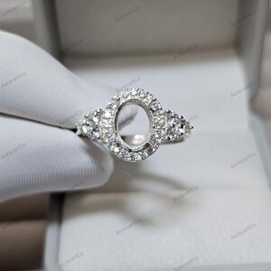 May include: A silver ring featuring an oval centre setting, encircled by a halo of small, sparkling stones. Further clusters of stones decorate each side of the ring, creating a dazzling display. The ring is set against a white background.