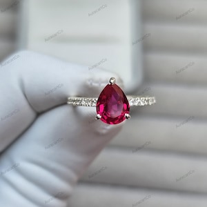 May include: A silver ring with a prominent, teardrop-shaped, deep red gemstone. The band is embellished with small, clear gemstones. The ring is displayed against a soft, neutral background.