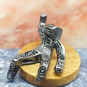 May include: A silver-coloured metal sculpture of a person in a relaxed pose, with intricate geometric patterns etched across its surface. The sculpture is mounted on a round, wooden base, showcasing a modern art aesthetic.