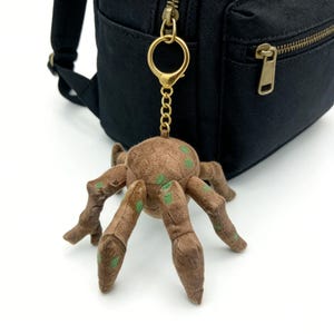 Op de afbeelding: Een bruine pluche spinkeychain met groene stippen en beweegbare poten hangt aan een goudkleurige sluiting die aan een zwarte rugzak is bevestigd. Het lichaam van de spin is rond en de poten zijn donkerder bruin.