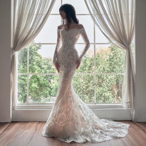 May include: A floor-length, off-the-shoulder gown featuring long sleeves and a fitted bodice. The dress is crafted from white lace with intricate floral patterns layered over a nude-toned fabric, creating a mermaid silhouette with a train.
