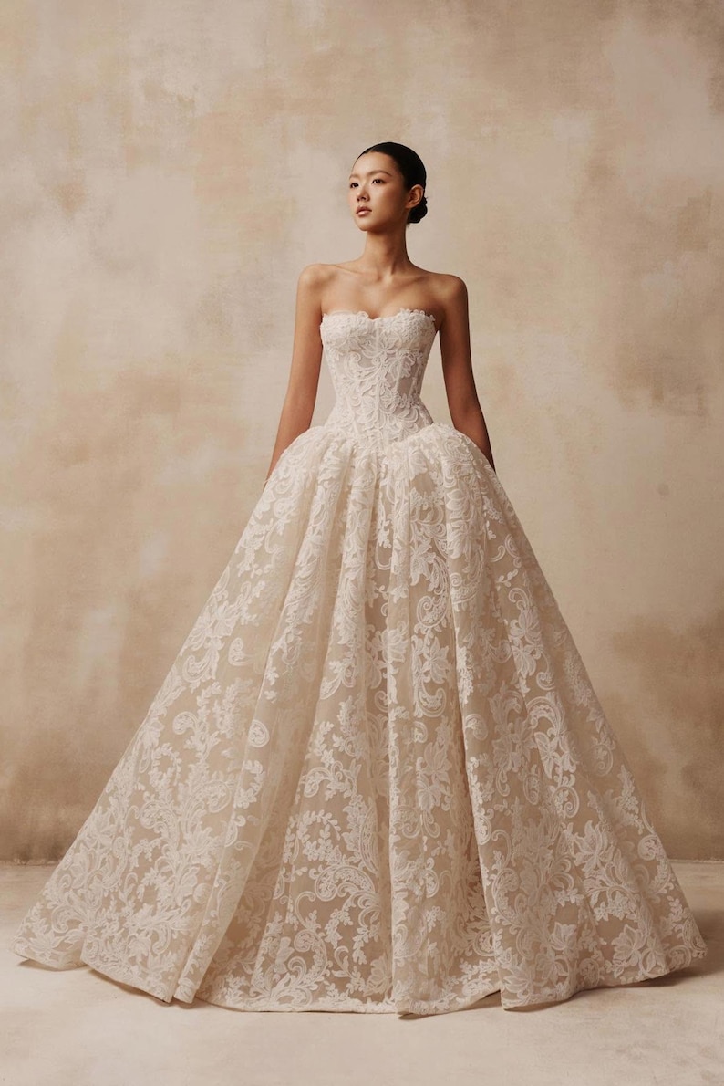 May include: A strapless floor-length ball gown featuring a fitted corset bodice and a voluminous skirt. The dress is crafted from a light beige fabric overlaid with intricate white floral lace patterns that extend from the bodice to the hem.