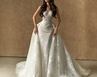 May include: A floor-length white strapless wedding dress featuring an intricate lace overlay, a fitted bodice with detailed embroidery, and a voluminous detachable overskirt that creates a dramatic silhouette with a long, flowing train.