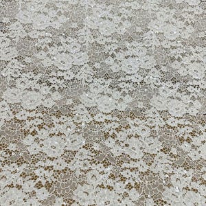 Peut inclure: Un tissu en dentelle blanche pr&eacute;sentant un motif floral dense avec des bords festonn&eacute;s complexes et de petites paillettes transparentes dispers&eacute;es dans tout le design, cr&eacute;ant un aspect d&eacute;licat et textur&eacute; sur toute la surface.