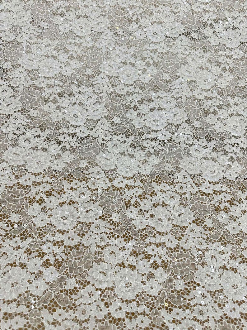 Peut inclure: Un tissu en dentelle blanche pr&eacute;sentant un motif floral dense avec des bords festonn&eacute;s complexes et de petites paillettes transparentes dispers&eacute;es dans tout le design, cr&eacute;ant un aspect d&eacute;licat et textur&eacute; sur toute la surface.
