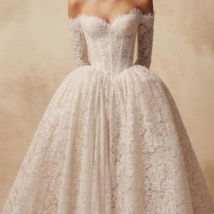 Peut inclure: Une robe blanche en dentelle &agrave; &eacute;paules d&eacute;nud&eacute;es avec des manches longues, un d&eacute;collet&eacute; en c&oelig;ur et un corsage structur&eacute; style corset qui se prolonge par une jupe volumineuse pleine longueur orn&eacute;e de motifs floraux complexes et de paillettes.