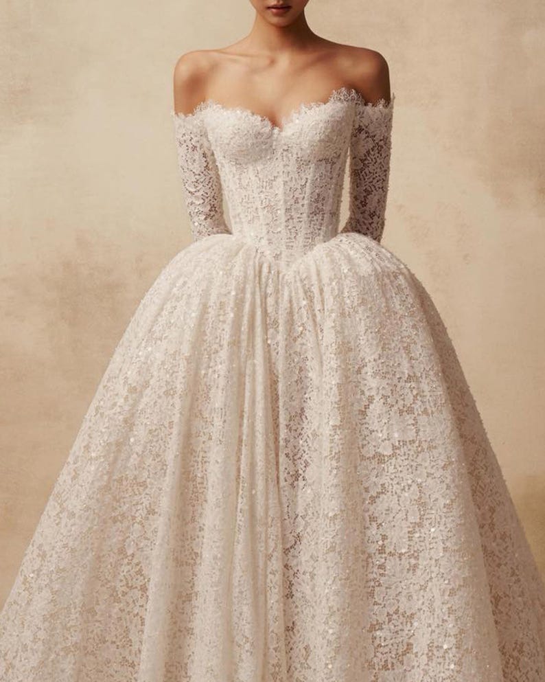 Peut inclure: Une robe blanche en dentelle &agrave; &eacute;paules d&eacute;nud&eacute;es avec des manches longues, un d&eacute;collet&eacute; en c&oelig;ur et un corsage structur&eacute; style corset qui se prolonge par une jupe volumineuse pleine longueur orn&eacute;e de motifs floraux complexes et de paillettes.