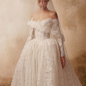 Peut inclure: Une robe de mari&eacute;e longue en dentelle blanche avec une encolure d&eacute;collet&eacute;e, des manches longues et un corsage structur&eacute;. La jupe volumineuse est confectionn&eacute;e en dentelle florale complexe avec des paillettes subtiles et un voile transparent.