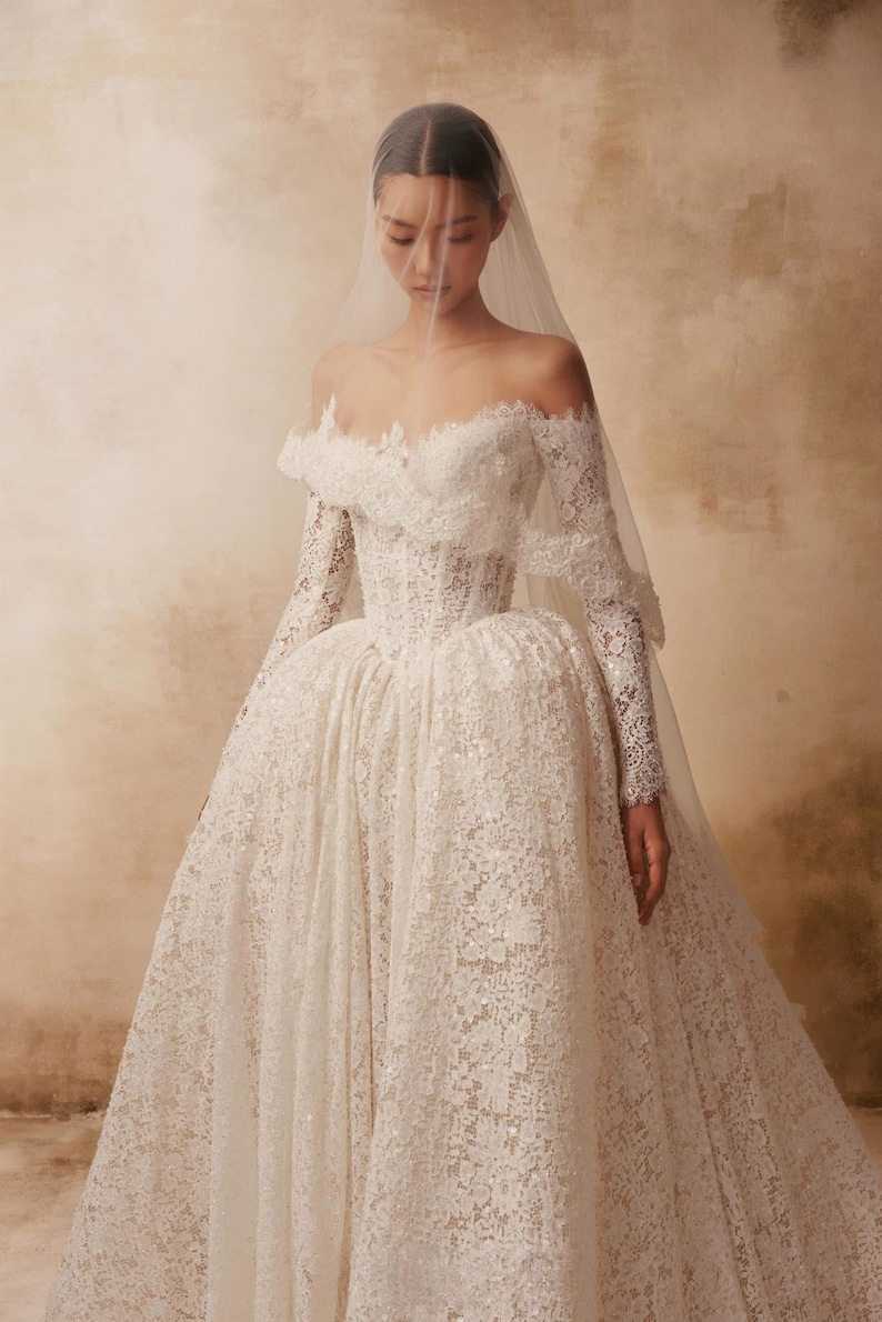 Peut inclure: Une robe de mari&eacute;e longue en dentelle blanche avec une encolure d&eacute;collet&eacute;e, des manches longues et un corsage structur&eacute;. La jupe volumineuse est confectionn&eacute;e en dentelle florale complexe avec des paillettes subtiles et un voile transparent.