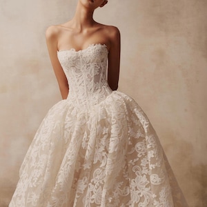 May include: A strapless ivory wedding dress featuring a fitted corset bodice and a voluminous, floor-length skirt. The entire garment is covered in intricate white floral lace embroidery layered over a light beige base fabric.