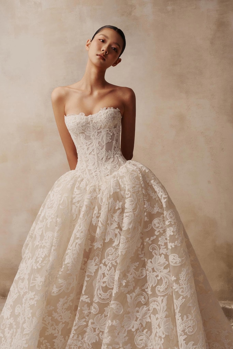 May include: A strapless ivory wedding dress featuring a fitted corset bodice and a voluminous, floor-length skirt. The entire garment is covered in intricate white floral lace embroidery layered over a light beige base fabric.