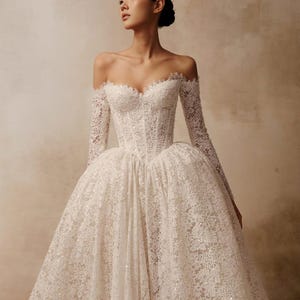 Peut inclure: Une robe longue en dentelle blanche avec une encolure en c&oelig;ur d&eacute;collet&eacute;e, des manches longues ajust&eacute;es et un corsage structur&eacute; style corset qui se prolonge par une jupe volumineuse orn&eacute;e de d&eacute;tails complexes en dentelle florale.