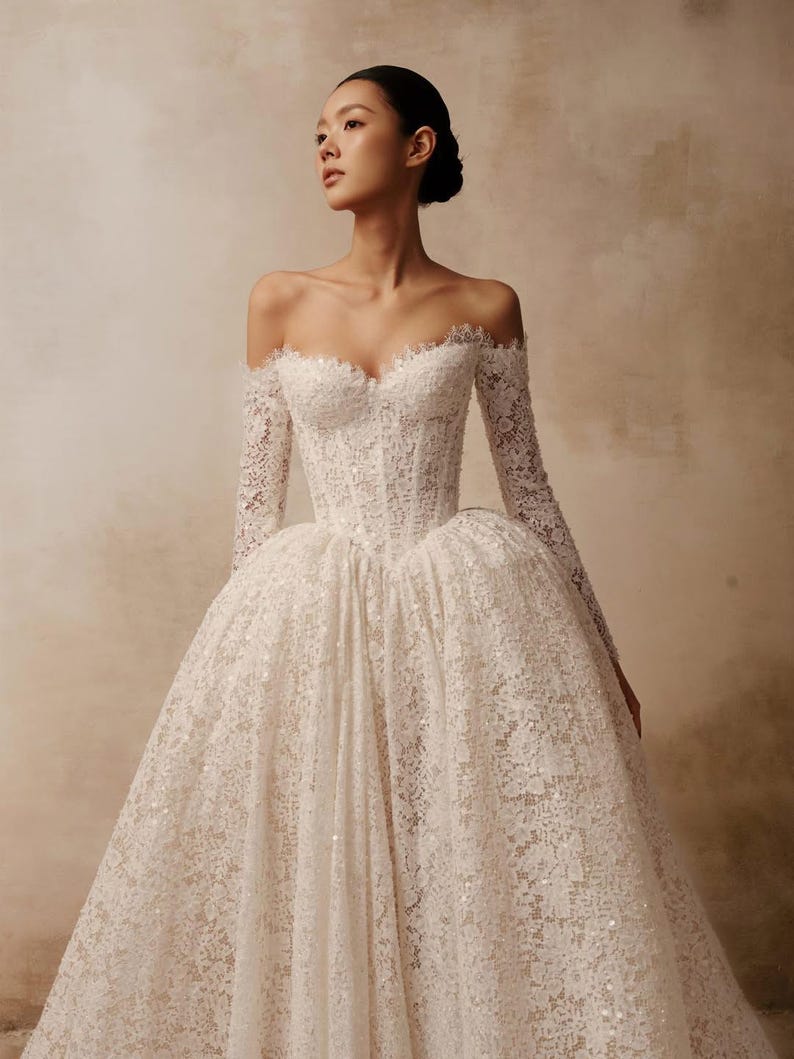 Peut inclure: Une robe longue en dentelle blanche avec une encolure en c&oelig;ur d&eacute;collet&eacute;e, des manches longues ajust&eacute;es et un corsage structur&eacute; style corset qui se prolonge par une jupe volumineuse orn&eacute;e de d&eacute;tails complexes en dentelle florale.